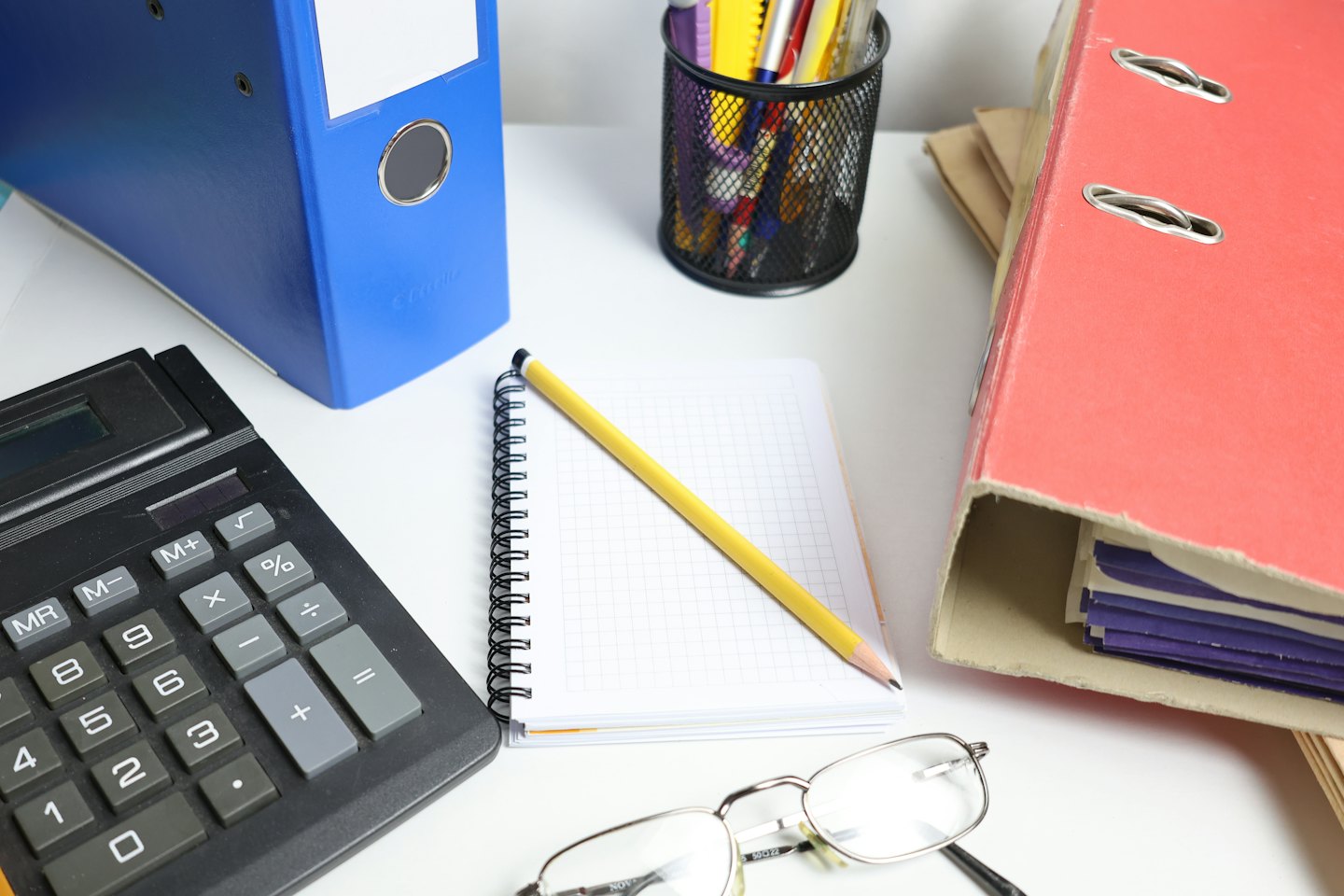 Schreibtisch mit Taschenrechner, Ordnern, Notizblock und Brille.